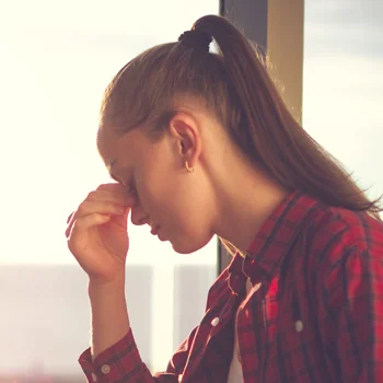 Eye Doctor Plainfield IL Glare and Contrast BOX A young woman pinching her nose in discomfort while the sun glares through a window behind her